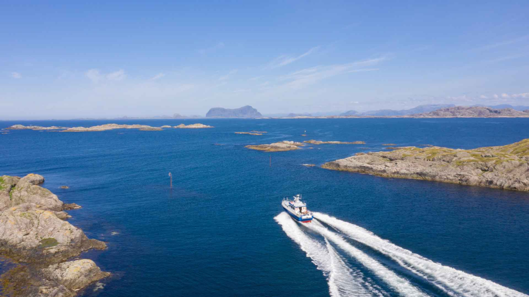Øyhopping med postbåten ein båt køyrer full fart mellom holmar og skjær, på veg ut mot det store hav. det er eit stort fjell i horisonten som skil seg ut då det er forma som ein hest. fleire fjell ligg i horisonten av bilde. båten er kvit, med blå detaljar. den skal til å køyre mellom to stakar.