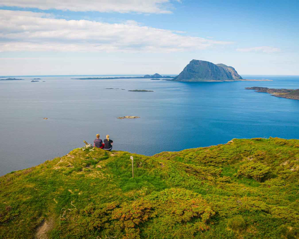 Atløy Brurastakken-4 to personer sitter på ein tupp på ein fjelltopp. nedanfor dei er ope hav. i enden av bilde står eit stort fjell. dei to damene sitt med ryggen til kameravinkelen.