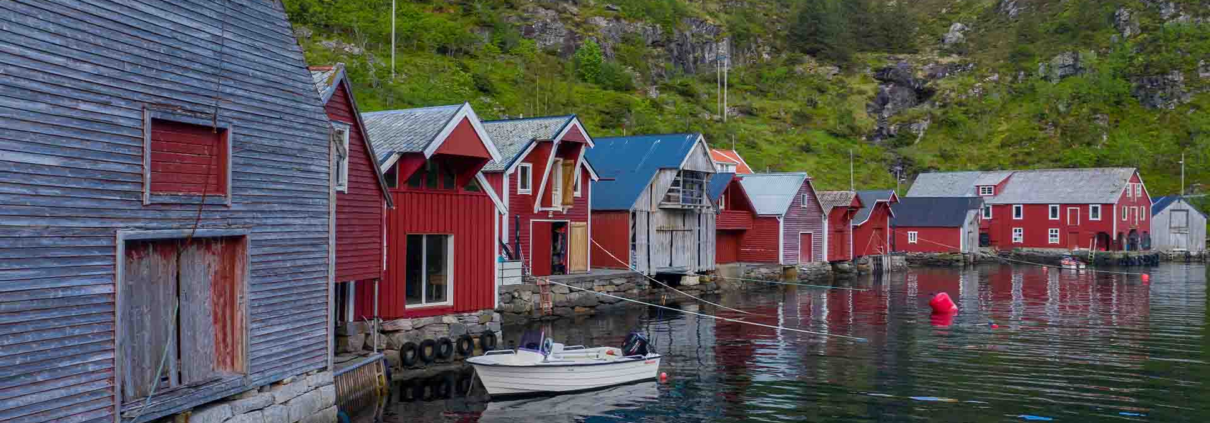 fleire naust står langs sjøen. nausta er raude og grå. ein båt ligg ved eine naustet. bakom nausta er det eit fjell med grønt gras.