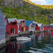 fleire naust står langs sjøen. nausta er raude og grå. ein båt ligg ved eine naustet. bakom nausta er det eit fjell med grønt gras.