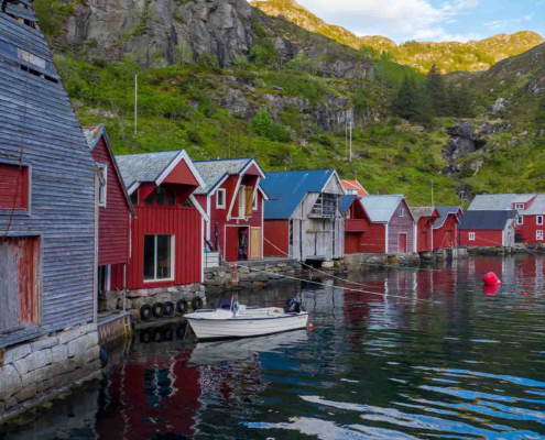 fleire naust står langs sjøen. nausta er raude og grå. ein båt ligg ved eine naustet. bakom nausta er det eit fjell med grønt gras.