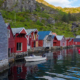 fleire naust står langs sjøen. nausta er raude og grå. ein båt ligg ved eine naustet. bakom nausta er det eit fjell med grønt gras.