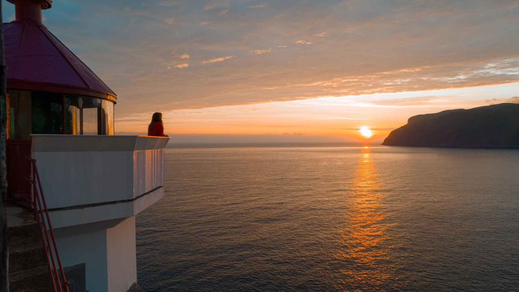 Hendanes Fyr, Måløy ei jente står ved eit fyrtårn og ser ut mot havet og solnedgangen. fyret er rødt med kvite detaljar. sola er nesten i havet, og solstrålene skin i havet.