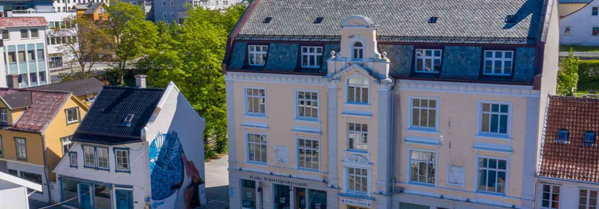 eit dronebilde av bygninger i Florø. eit stort, rosa gamalt bygg står i front av bilde. ein person syklar med vogn nede i gata. det er blå himmel