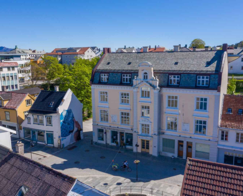 eit dronebilde av bygninger i Florø. eit stort, rosa gamalt bygg står i front av bilde. ein person syklar med vogn nede i gata. det er blå himmel