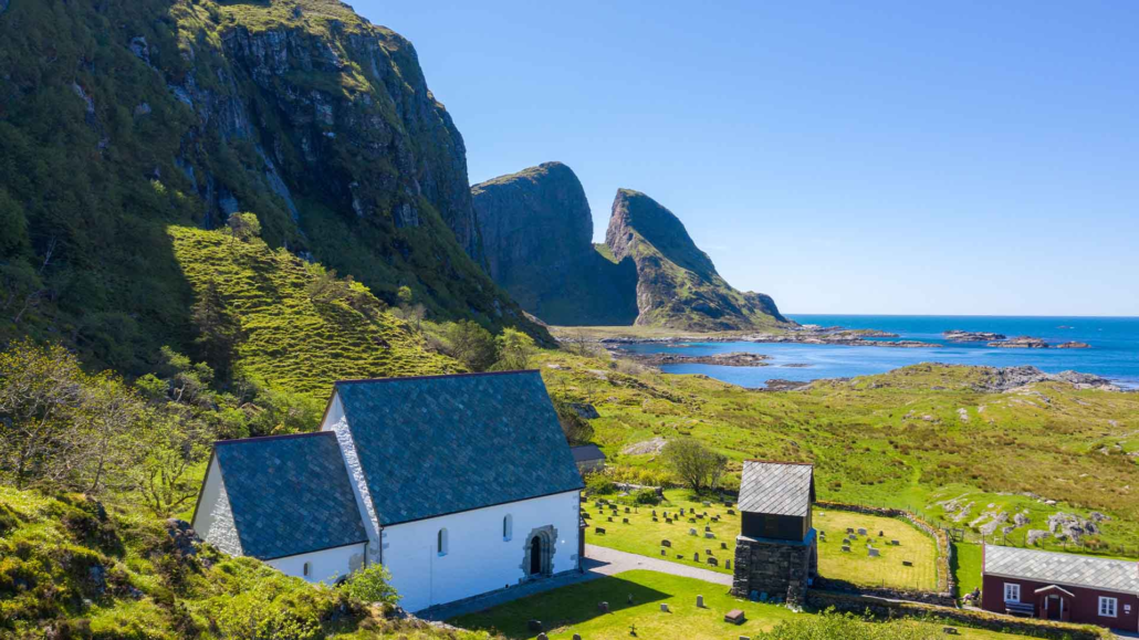 SAGAØYA KINN - FOTO VISIT FJORDKYSTEN grønt gras og blå himmel i omgivelsene. i front av bilde står ei steinkyrkje vendt mot havet. foran kyrkja er det graver. det er eit lite raudt hus på høgre side av kirka. eit fjell stikk seg ut bakom kirka. Kinnaklova, som har to klover som står mot kvarandre viser seg mektig mot bilde.