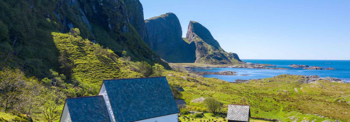 grønt gras og blå himmel i omgivelsene. i front av bilde står ei steinkyrkje vendt mot havet. foran kyrkja er det graver. det er eit lite raudt hus på høgre side av kirka. eit fjell stikk seg ut bakom kirka. Kinnaklova, som har to klover som står mot kvarandre viser seg mektig mot bilde.