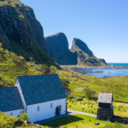 grønt gras og blå himmel i omgivelsene. i front av bilde står ei steinkyrkje vendt mot havet. foran kyrkja er det graver. det er eit lite raudt hus på høgre side av kirka. eit fjell stikk seg ut bakom kirka. Kinnaklova, som har to klover som står mot kvarandre viser seg mektig mot bilde.