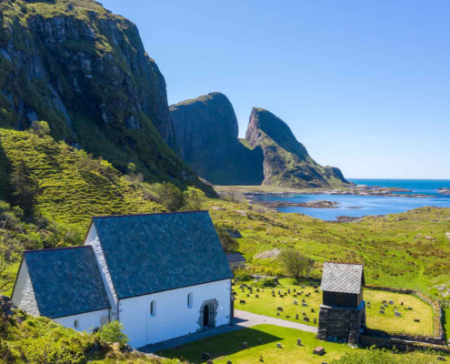 grønt gras og blå himmel i omgivelsene. i front av bilde står ei steinkyrkje vendt mot havet. foran kyrkja er det graver. det er eit lite raudt hus på høgre side av kirka. eit fjell stikk seg ut bakom kirka. Kinnaklova, som har to klover som står mot kvarandre viser seg mektig mot bilde.